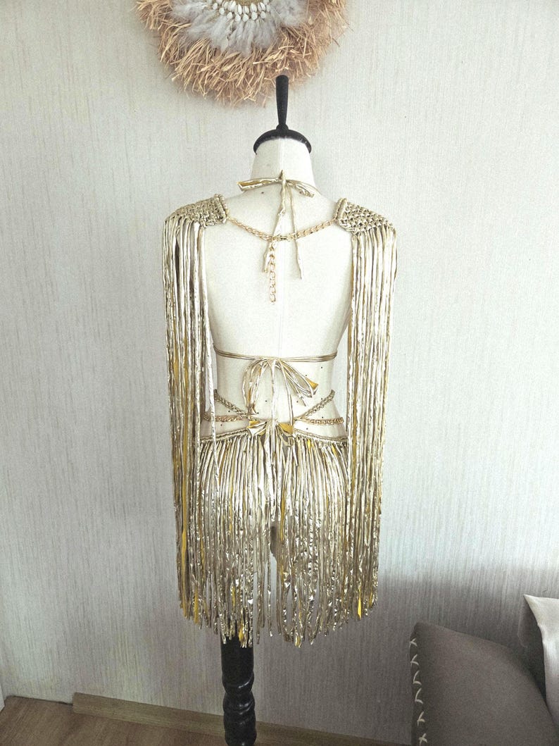 May include: A metallic gold fringe garment displayed on a mannequin, featuring long shoulder fringe panels, a back tie closure, and a waist section with multiple thin straps and dangling fringe elements that extend down to the lower back area.