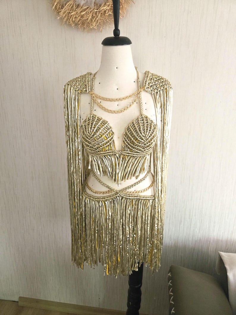 May include: A metallic gold two-piece outfit featuring a structured bralette top and a matching skirt, both adorned with long, shimmering fringe. The set includes integrated gold chain accents at the neckline and waist, with shoulder capes.