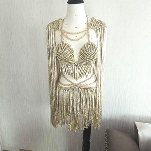 May include: A metallic gold two-piece outfit on a mannequin, featuring a shell-shaped bra top and a matching skirt, both adorned with long, flowing fringe. The set includes gold chain accents at the neckline and waist, with integrated shoulder capes.