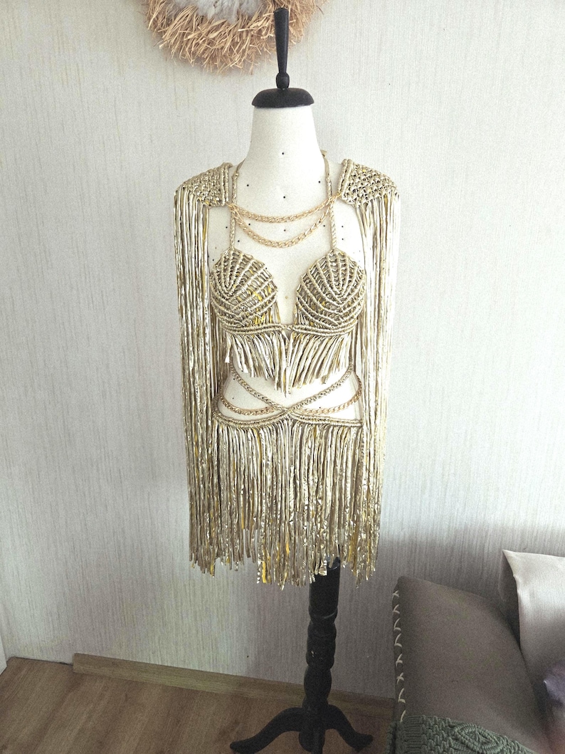 May include: A metallic gold two-piece outfit on a mannequin, featuring a shell-shaped bra top and a matching skirt, both adorned with long, flowing fringe. The set includes gold chain accents at the neckline and waist, with integrated shoulder capes.