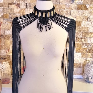 Op de afbeelding: Een zwarte macramé choker met gouden accenten en lange franjes. De ketting wordt gedragen door een wit mannequin.