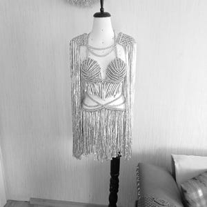 May include: A silver two-piece outfit on a mannequin, featuring a structured bra top and a matching skirt, both adorned with long, shimmering fringe. The set includes integrated shoulder capes and delicate chain-like detailing across the chest and waist.