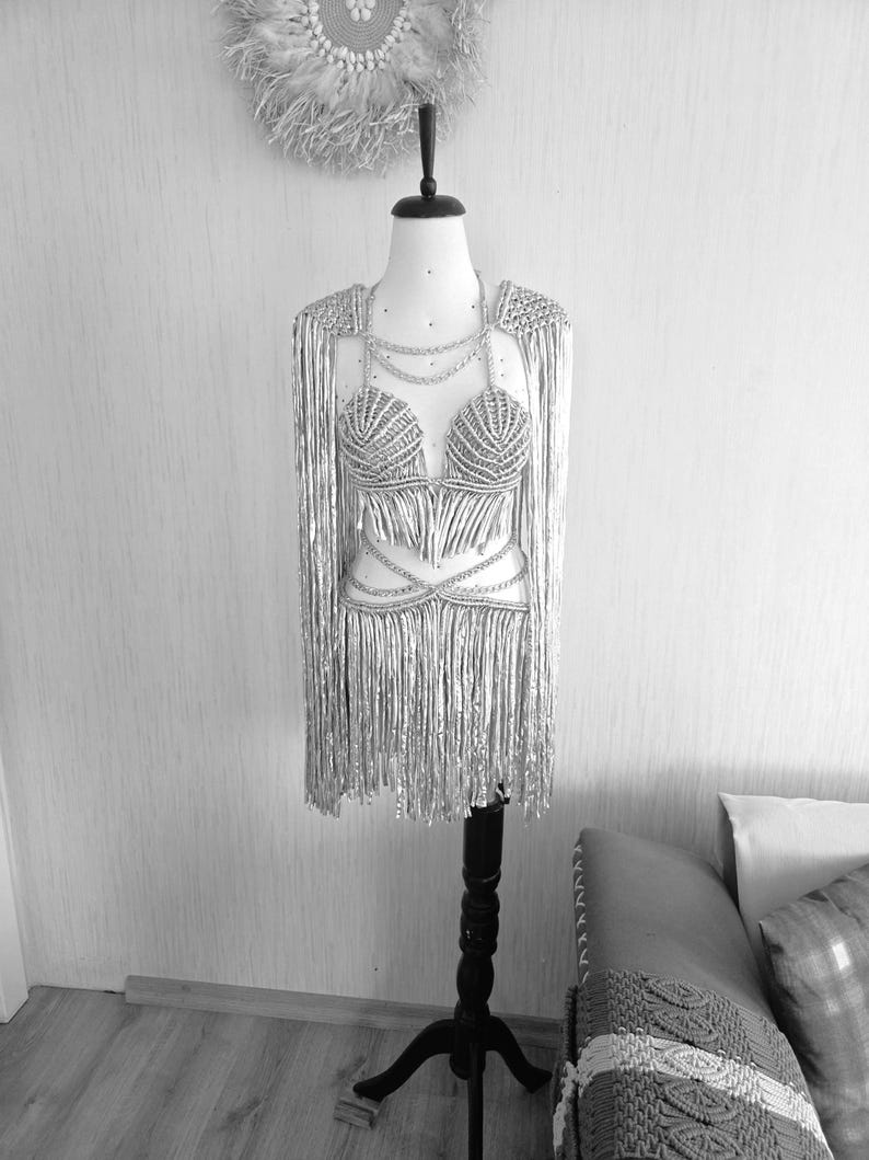 May include: A silver two-piece outfit on a mannequin, featuring a structured bra top and a matching skirt, both adorned with long, shimmering fringe. The set includes integrated shoulder capes and delicate chain-like detailing across the chest and waist.