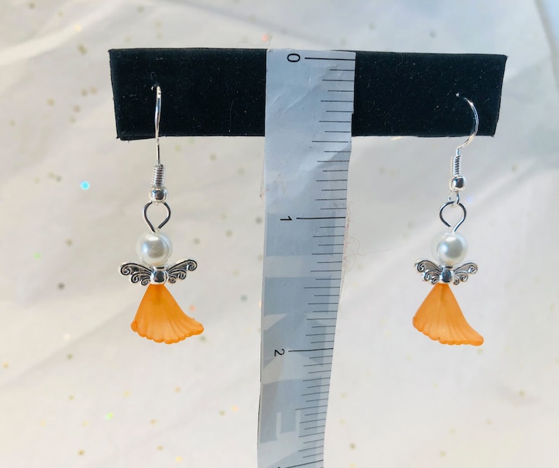 Puede incluir: Un par de pendientes de &aacute;ngel de plata con alas naranjas y una perla blanca. Los pendientes miden aproximadamente 2,5 cm de largo.