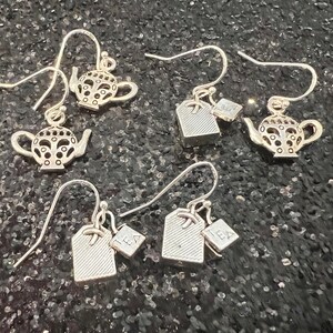 May include: Four pairs of silver earrings with a teapot and tea bag charm. The tea bag charms have the word "tea" on them.