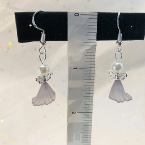 Puede incluir: Un par de pendientes de &aacute;ngel de plata con una perla blanca y un dije en forma de flor lavanda. Los pendientes miden aproximadamente 2,5 cm de largo.