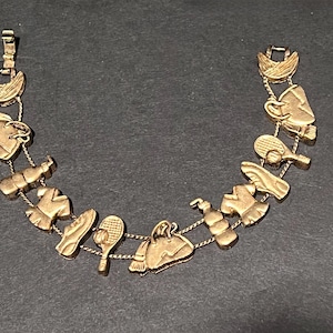 May include: Gold-toned charm bracelet featuring sports and travel-themed charms. The bracelet includes charms shaped like a tennis racket, a handbag, a shoe, a shirt, and a coffee cup. The bracelet is made of metal and has a clasp closure.