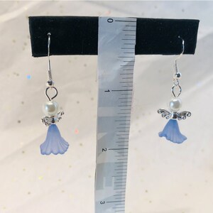 Puede incluir: Un par de pendientes de plata con forma de &aacute;ngel, con una flor azul y una perla blanca.