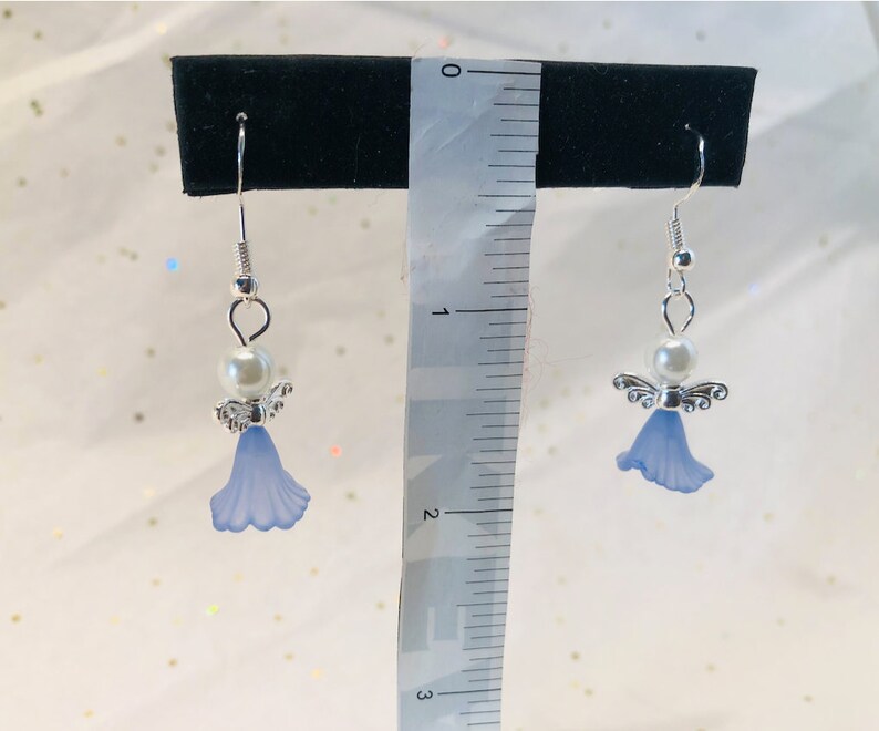 Puede incluir: Un par de pendientes de plata con forma de &aacute;ngel, con una flor azul y una perla blanca.