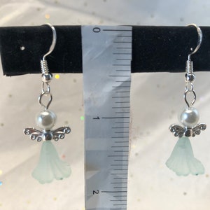 Puede incluir: Un par de pendientes de &aacute;ngel de plata con un &aacute;ngel de cristal azul claro y una perla blanca. Los pendientes miden aproximadamente 2,5 cm de largo.