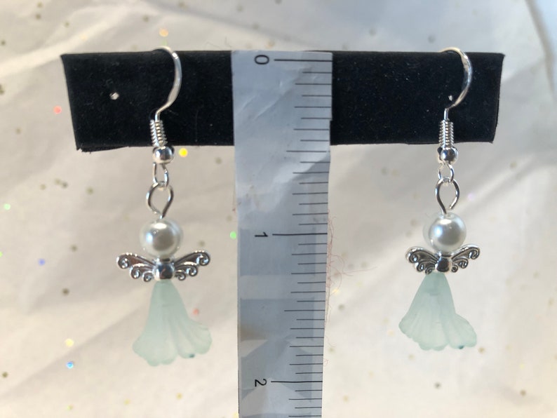 Puede incluir: Un par de pendientes de &aacute;ngel de plata con un &aacute;ngel de cristal azul claro y una perla blanca. Los pendientes miden aproximadamente 2,5 cm de largo.