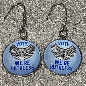 May include: A pair of silver dangle earrings with blue and white round charms. The charms feature a black silhouette of a collar and the text "VOTE WE'RE RUTHLESS".