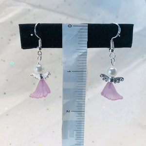 Puede incluir: Un par de pendientes de plata con forma de &aacute;ngel con una perla y un dije de ala de &aacute;ngel de color lavanda. Los pendientes est&aacute;n colgados de una regla que mide 5 cm.