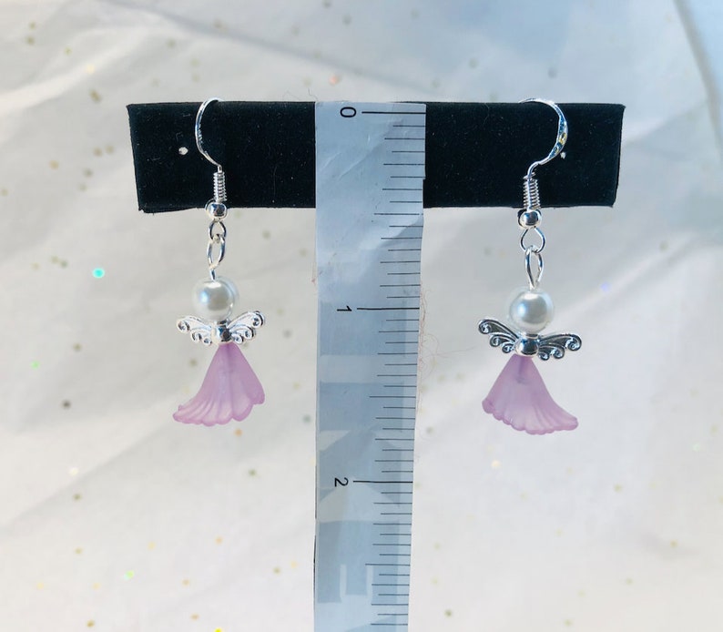 Puede incluir: Un par de pendientes de plata con forma de &aacute;ngel con una perla y un dije de ala de &aacute;ngel de color lavanda. Los pendientes est&aacute;n colgados de una regla que mide 5 cm.