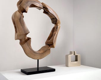 Op de afbeelding: Een grote, abstracte houten sculptuur met een cirkelvormig ontwerp, gemonteerd op een zwarte rechthoekige basis. De sculptuur heeft een lichtbruine kleur en een unieke, organische vorm. Een kleine, beige keramische vaas met een vierkante opening staat in de buurt.