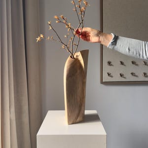 Può includere: Un vaso in legno alto e slanciato con una venatura liscia color marrone chiaro e un'apertura superiore curva e asimmetrica. Il vaso presenta una forma verticale scultorea e contiene un ramo sottile con piccoli e delicati fiori color arancio chiaro.