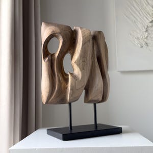 Può includere: Una scultura in legno con finitura in legno naturale, caratterizzata da forme organiche e due aperture circolari. La scultura è montata su un supporto in metallo nero con base rettangolare, esposta su una superficie bianca.