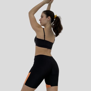 K&ouml;nnte beinhalten: Eine Frau tr&auml;gt einen schwarzen Sport-BH und schwarze Radlerhosen mit orangefarbenen Akzenten. Die Shorts haben einen hohen Bund und eine enge Passform.