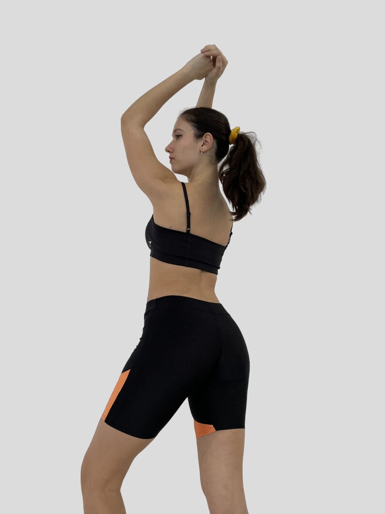 K&ouml;nnte beinhalten: Eine Frau tr&auml;gt einen schwarzen Sport-BH und schwarze Radlerhosen mit orangefarbenen Akzenten. Die Shorts haben einen hohen Bund und eine enge Passform.
