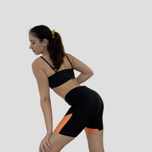 K&ouml;nnte beinhalten: Eine Frau tr&auml;gt einen schwarzen Sport-BH und schwarze und orangefarbene Radshorts. Die Shorts haben ein kontrastierendes orangefarbenes Panel an der Seite.