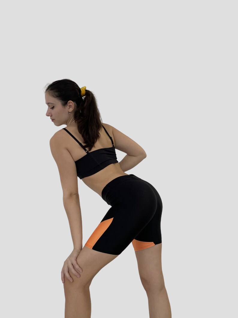 K&ouml;nnte beinhalten: Eine Frau tr&auml;gt einen schwarzen Sport-BH und schwarze und orangefarbene Radshorts. Die Shorts haben ein kontrastierendes orangefarbenes Panel an der Seite.