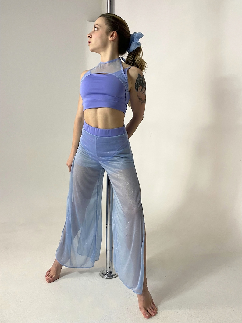 May include: A light blue mesh top and matching pants with a high waist and side slits. The top has a halter neck and a mesh panel across the chest. The outfit is worn by a person standing next to a silver pole.