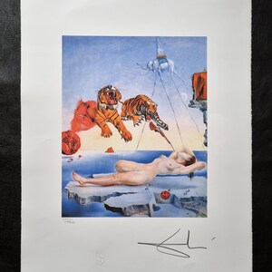 May include: A surrealist print featuring a nude figure on an ice floe, with tigers, an elephant, and pomegranates. The artwork has a blue and orange color palette, with a signature in the lower right corner. The print is on a white paper.
