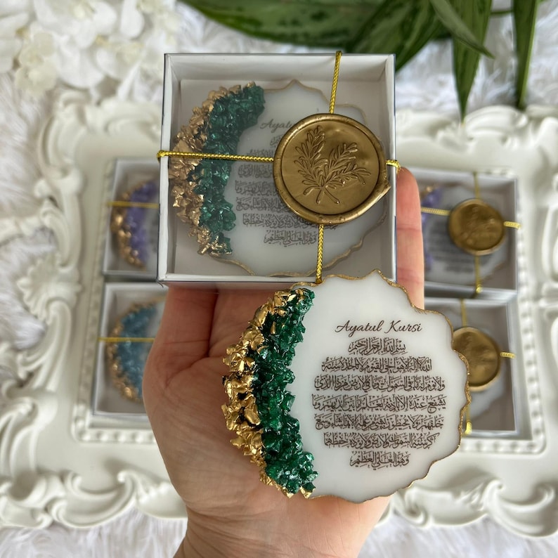 K&ouml;nnte beinhalten: Ein wei&szlig;es Harzornament mit gewelltem Rand, das auf einer Seite gr&uuml;ne und goldene strukturierte Akzente sowie den in Schwarz gedruckten arabischen Text von Ayatul Kursi aufweist. Das Ornament ist mit einer Goldschnur und einem goldenen Wachssiegel versehen.