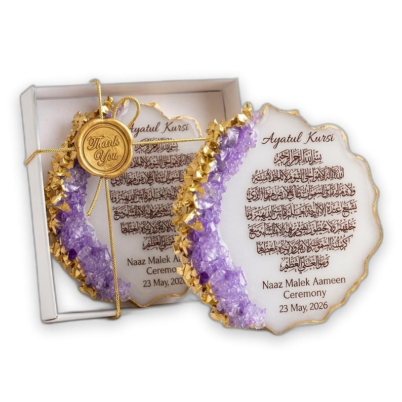 K&ouml;nnte beinhalten: Zwei wei&szlig;e, unregelm&auml;&szlig;ige Untersetzer im Geoden-Stil mit goldfarbenen R&auml;ndern und violetten Kristallakzenten. Sie zeigen arabische Kalligrafie des Ayatul Kursi, den Text 'Naaz Malek Aameen Ceremony 23 May, 2026' und ein goldenes Wachssiegel 'Thank You'.