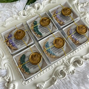 K&ouml;nnte beinhalten: Sechs quadratische Geschenkboxen mit halbmondf&ouml;rmigen Harzornamenten, verziert mit Blattgoldr&auml;ndern und eingebetteten violetten, gr&uuml;nen und blauen Kristallen. Jede Box ist mit einer goldenen Schnur und einem runden goldenen Wachssiegel versehen.