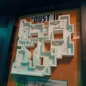 Puede incluir: Un modelo 3D enmarcado del mapa "Dust II", un mapa de desactivación de bombas. El modelo es blanco y está sobre un fondo naranja. El texto "DUST II", "LOCATION: MOROCCO", "TYPE: BOMB DEFUSAL" y "! RUSH B" son visibles.