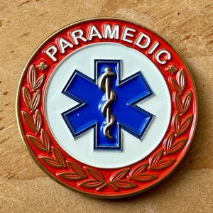 May include: A circular paramedic emblem in red, white, and blue. It features a blue star of life symbol with a silver caduceus, encircled by the word "PARAMEDIC". A gold laurel wreath surrounds the emblem. The emblem is a symbol of emergency medical services.