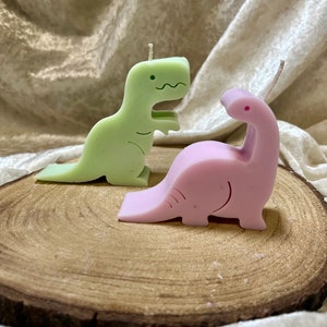 Könnte beinhalten: Zwei dinosaurierförmige Kerzen, eine grüne und eine rosa, auf einer Holzoberfläche. Die Kerzen sind so gestaltet, dass sie wie Dinosaurier aussehen, mit einem einfachen, cartoonartigen Stil.