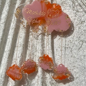 Könnte beinhalten: Ein rosafarbenes und orangefarbenes, wolkenförmiges Harz-Ornament mit goldenen Akzenten und den Namen "Mathilda Sophie" in goldenen Lettern. Das Ornament hängt an einer goldenen Kette und hat vier kleinere, wolkenförmige Ornamente, die darunter hängen.