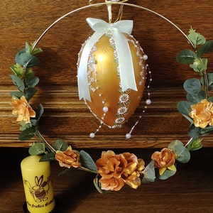 Könnte beinhalten: Ein goldenes Osterei mit weißen Perlenakzenten und einem hellgrünen Band hängt an einem goldenen Metallkranz, der mit orangefarbenen Blumen und grünen Blättern geschmückt ist.