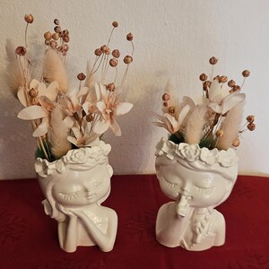 Könnte beinhalten: Zwei weiße Keramik-Blumentöpfe in Form eines Mädchenkopfes mit einer Blumenkrone. Jeder Topf ist mit getrockneten Blumen und Grün gefüllt.