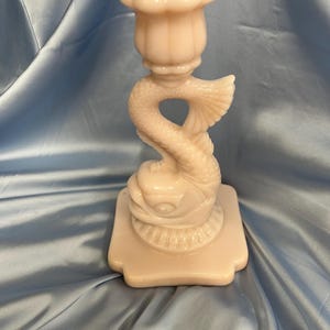 May include: A pale pink glass candlestick holder. The candlestick features a dolphin design with a textured body and a flared base. The top of the candlestick has a flower-shaped cup to hold a candle. The candlestick is made of glass.