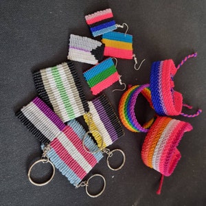 Puede incluir: Una colección de pulseras y llaveros de cuentas de colores que representan varias banderas del orgullo, incluidas las banderas transgénero, bisexual y pansexual. Las pulseras están hechas con una variedad de colores y patrones, y los llaveros están unidos a llaveros de plata.