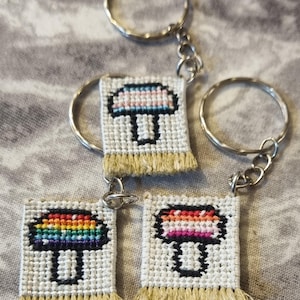 May include: Three handmade keychains with embroidered mushroom designs. Each keychain features a white background with a colorful mushroom and a fringe at the bottom. The keychains are attached to silver key rings.