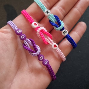Puede incluir: Cuatro pulseras de la amistad con diferentes colores y cuentas. Una pulsera es morada con cuentas blancas que dicen "3005". Otra pulsera es rosa con cuentas blancas en forma de corazón. Una tercera pulsera es azul con cuentas blancas que dicen "KE". La última pulsera es azul claro con una cuenta blanca que dice "K".