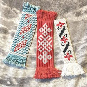 May include: Three handmade bookmarks with fringe. The bookmark on the left is blue and white with a geometric pattern. The bookmark in the middle is red and white with a knot pattern. The bookmark on the right is white with black and red floral designs.