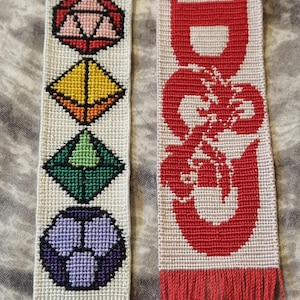 May include: Two white fabric bookmarks with red fringe. The bookmark on the left has a white background with four colorful geometric shapes. The bookmark on the right has a white background with a red dragon design and the text "D&D".