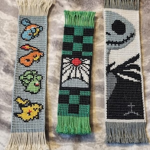 Può includere: Tre segnalibri fatti a mano con perline di diversi design. Il primo segnalibro è blu e bianco con un personaggio di Pokemon, il secondo è verde e nero con un motivo a scacchi e un fiore bianco con un centro rosso, e il terzo è grigio e nero con un personaggio dei cartoni animati con un ampio sorriso.