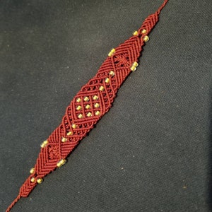 May include: A maroon macrame bracelet with a geometric pattern and small yellow beads.