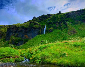 Könnte beinhalten: Ein Wasserfall stürzt einen üppigen grünen Hang hinunter, im Vordergrund fließt ein kleiner Bach. Der Himmel ist eine Mischung aus Blau und Grau mit fluffigen Wolken.