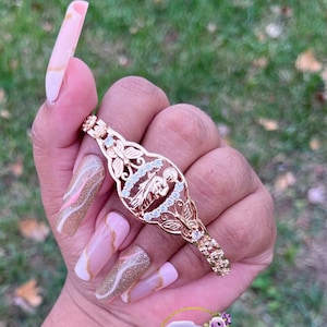 May include: Gold bracelet with a detailed design featuring a religious figure and butterflies. The bracelet is on a person's wrist.