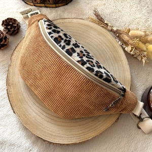 Puede incluir: Un cinturón de pana marrón con un panel con estampado de leopardo y una correa beige. La bolsa tiene una cremallera y herrajes dorados. El cinturón está sobre una superficie de madera.