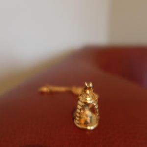 May include: A gold-colored pendant necklace featuring a small, ornate, cage-like charm. The charm has a detailed design and is suspended from a delicate chain. The necklace is displayed on a red surface.
