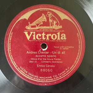 May include: A vintage Victrola record with a red label featuring the text "Victrola" and "His Master's Voice." The label includes the title "Andrea Chenier - Un di all azzurro spazio" by Enrico Caruso. The record is black.