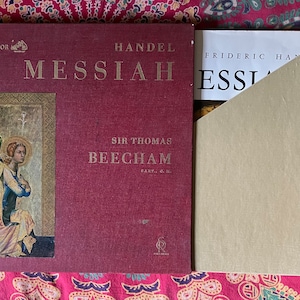May include: A red vinyl record album with a gold angel illustration on the front cover. The album is titled "Handel Messiah" and features the name "Sir Thomas Beecham" in gold lettering. The album is part of the RCA Victor collection.